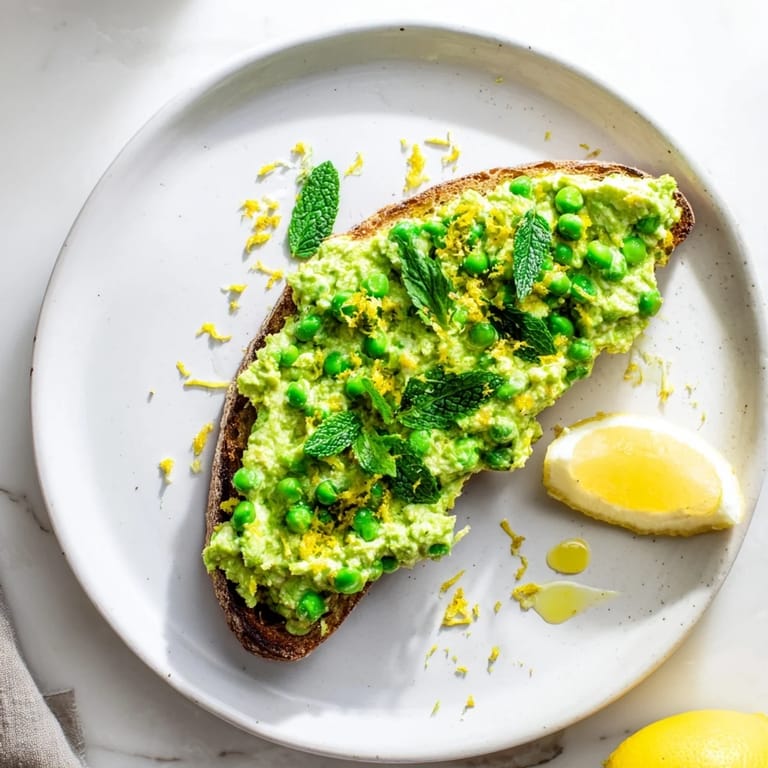 A close view of Pea & Mint Ricotta Toast reveals creamy whipped ricotta dotted with tender peas and chopped mint on crisp, toasted sourdough.