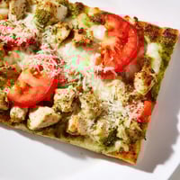 A close-up of a freshly baked Chicken Pesto Pizza with melted mozzarella and bright tomato slices, garnished with fresh basil on a wooden board.